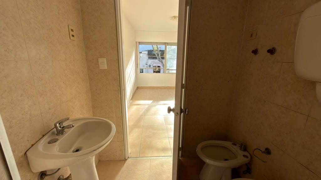 # Bathroom Description
This bathroom features beige ceramic tile walls and flooring throughout, with two separate toilet fixtures visible in the space. The room includes a wall-mounted sink with chrome fixtures on the left side and appears to be a dual or split bathroom layout. Natural light streams in through a doorway leading to an adjacent room with windows, and the space shows basic finishes in fair condition with functional plumbing fixtures.