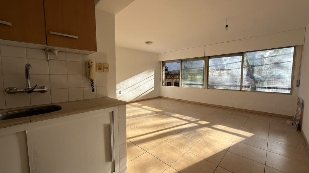 # Property Description
This is a bright, open-plan kitchen and living area featuring cream-colored tile flooring and abundant natural light from large horizontal windows with views to the exterior. The kitchen includes white subway tile backsplash, brown upper cabinetry, and stainless steel appliances, while the spacious living area offers excellent proportions and a modern, minimalist aesthetic. The room is in good condition with clean finishes and appears to be a contemporary residential space with excellent natural illumination throughout.