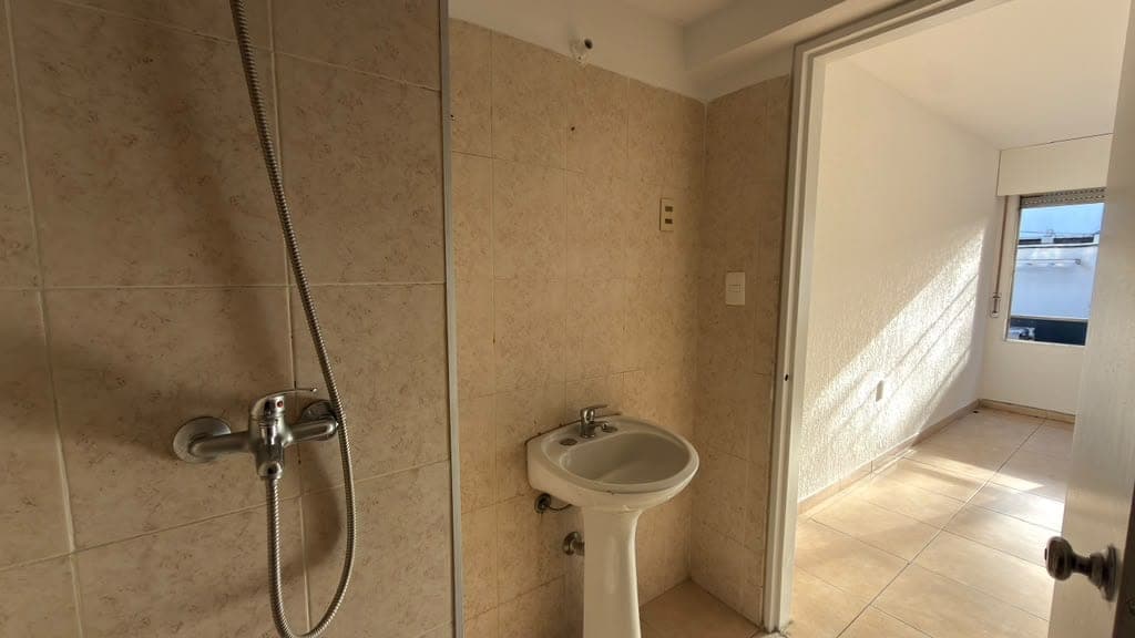 # Bathroom Description
This is a spacious bathroom featuring beige/tan marble-look tile walls and light tile flooring throughout. The room includes a pedestal sink, a walk-in shower with glass enclosure and chrome fixtures, and bright natural light from a window. The bathroom is in good condition with clean finishes and modern plumbing fixtures, offering a bright and airy layout with access to an adjacent room visible through the doorway.