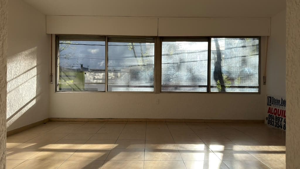 # Property Image Description
This is a bright, empty commercial or residential space featuring light beige walls and neutral tile flooring. The room is flooded with natural light from four large sliding glass windows with screens that offer views of the surrounding neighborhood and sky. The space appears clean and well-maintained, with modern window treatments and ample natural ventilation.
WATERMARK: Oficina Jalk