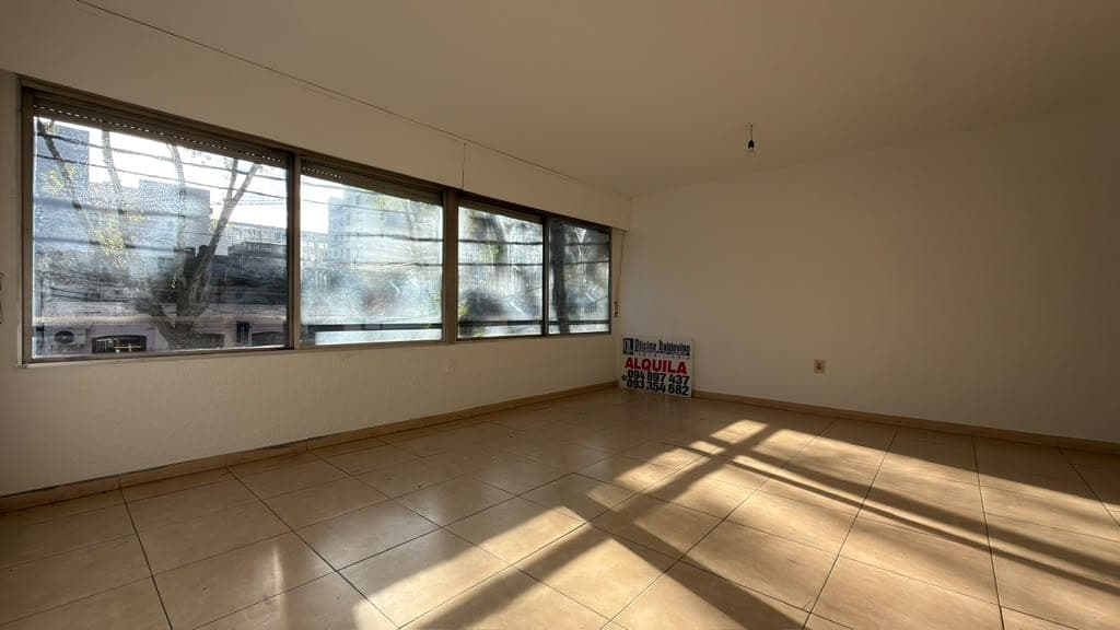 # Property Description
This is a bright, empty commercial or residential space featuring light beige tile flooring and neutral cream-colored walls. The room is flooded with natural light from a series of four large horizontal windows along one wall, offering views of the surrounding urban area and bare trees outside. The space appears clean and well-maintained, with minimal fixtures visible, making it ideal for retail, office, or residential use.
WATERMARK: Bossa Imobiliária