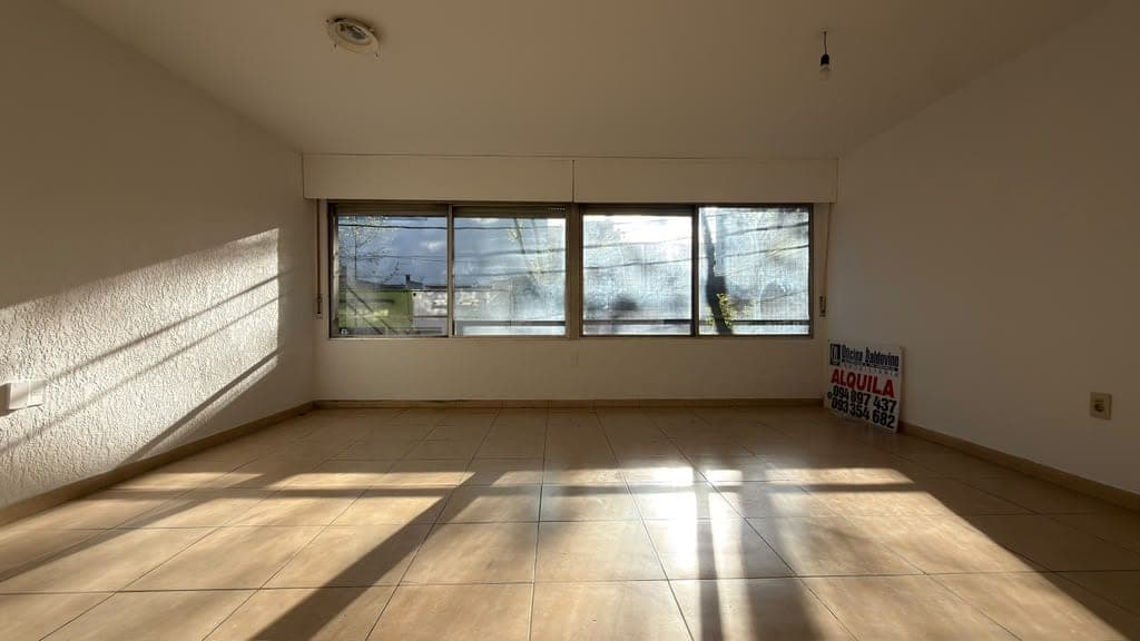 # Property Description
This is a bright, spacious commercial or residential space featuring light beige walls and neutral tile flooring. The room is flooded with natural light from four large horizontal windows that offer street-level views and create striking shadow patterns across the floor. The clean, minimalist interior with recessed ceiling lighting and simple wall outlets suggests this is an empty retail or office space ready for tenant occupancy.
WATERMARK: Inmobiliaria (rental/real estate agency sign visible on right wall)