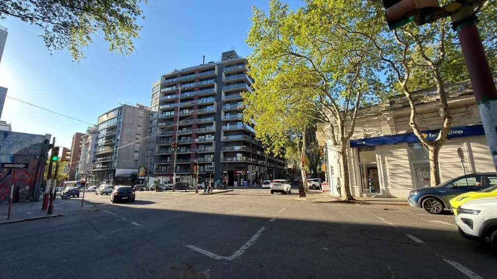 # Urban Street and Residential Complex
This image shows a vibrant urban streetscape featuring a modern multi-story residential building with contemporary balconies and mixed-use ground-floor retail spaces. The scene captures a tree-lined pedestrian area with mature plane trees providing shade, surrounded by additional mid-rise apartment buildings and street-level commercial storefronts, indicating a well-established urban neighborhood with good walkability and amenities.