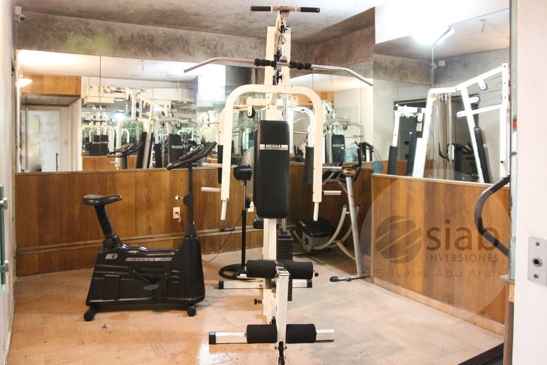This image shows a residential building's fitness room with mirrored walls, wood-paneled lower walls, and light tile-style flooring. Equipment includes a black Embreex 359 upright exercise bike, a white Megga multi-station home gym with a lat pulldown bar and leg roller, and a white cable station visible in the mirror reflections. The room appears functional but dated, with a textured gray ceiling and fluorescent lighting.
WATERMARK: SIAB Inversiones (Silveira Abu Arab)