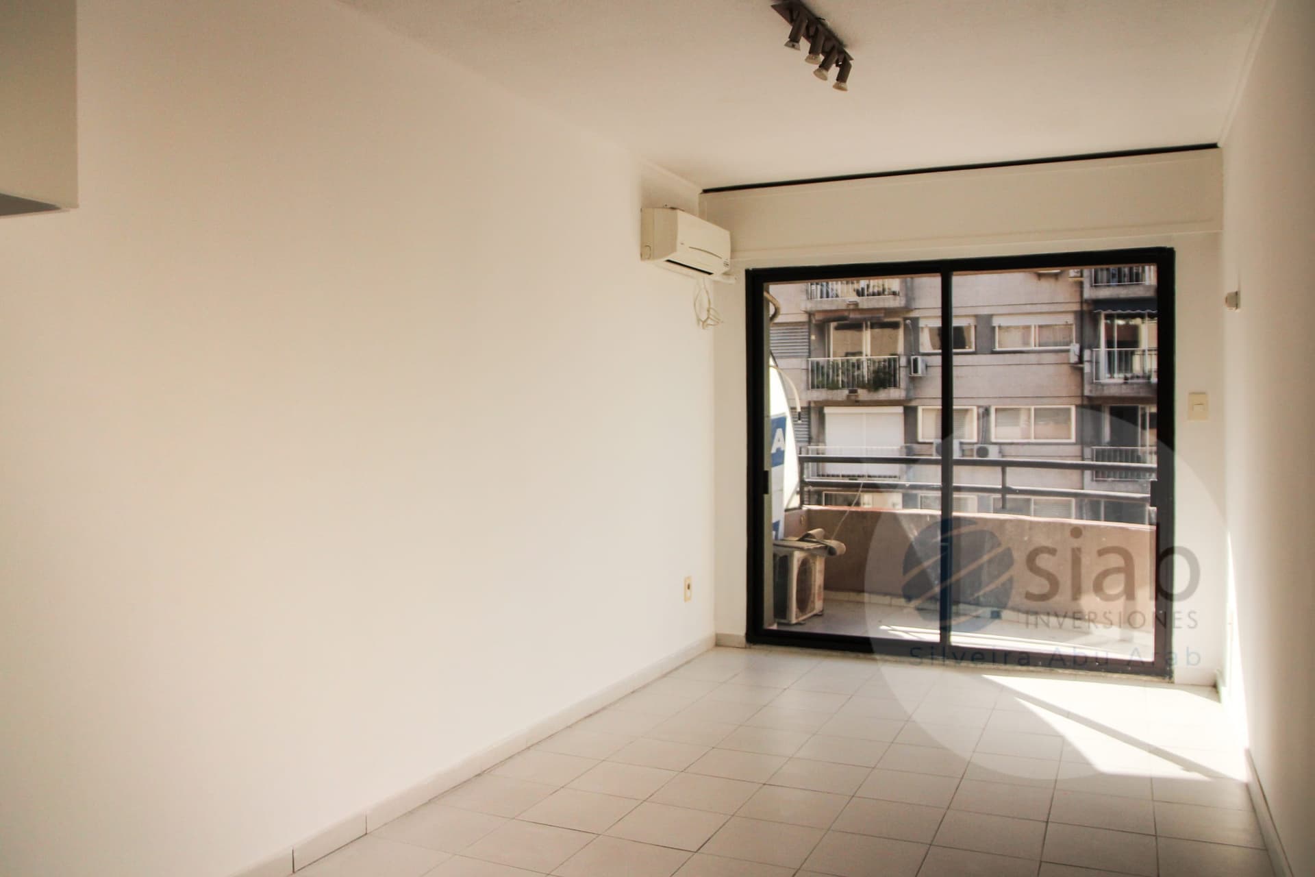 This is an empty living room or multipurpose space with light cream walls and large white ceramic floor tiles. A wall-mounted split air conditioning unit and a track-style ceiling spotlight fixture are visible, and a black-framed sliding glass door opens onto a balcony with a view of neighboring apartment buildings. The space is bright with natural light, and the room appears clean and unfurnished.
WATERMARK: Siao Inversiones