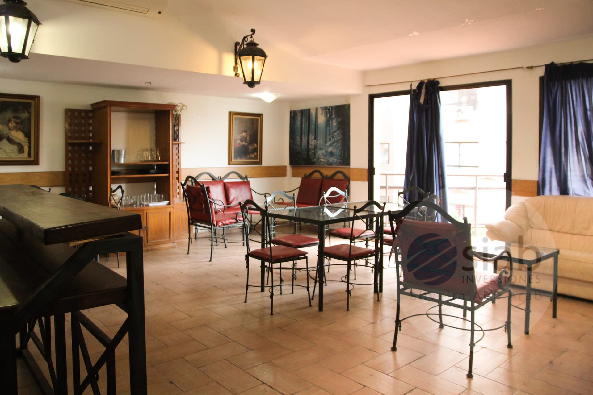 This is a spacious open-plan living and dining area with terracotta-tile flooring, wall-mounted and hanging lantern-style lights, and a wall-mounted air conditioning unit. The space includes a glass-top dining table with wrought-iron chairs upholstered in red, a red leather sofa and matching seating, a cream leather sofa, and a wooden display cabinet with glassware. Large sliding glass doors with navy drapes open onto a balcony, providing natural light.
WATERMARK: Siab Inversiones