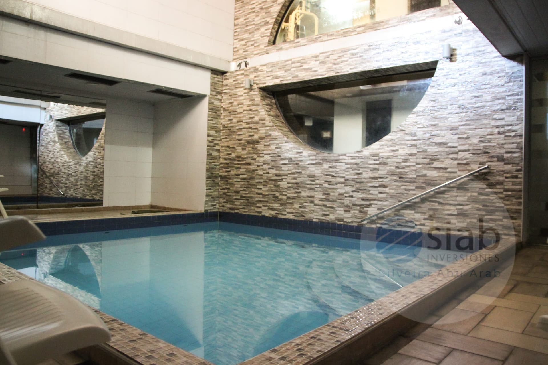 This image shows an indoor swimming pool with clear blue water, bordered by a mosaic tile edge and a dark blue tile band at the waterline. The adjacent walls feature textured stone-look tiles in grey and beige tones, with curved, arched cutouts and glass panels, along with a stainless steel handrail and a white lounge chair in the foreground. Warm wall lighting and a tiled deck give the space a spa-like, well-maintained appearance.
WATERMARK: Siab Inversiones (Silvia Abu Arab)