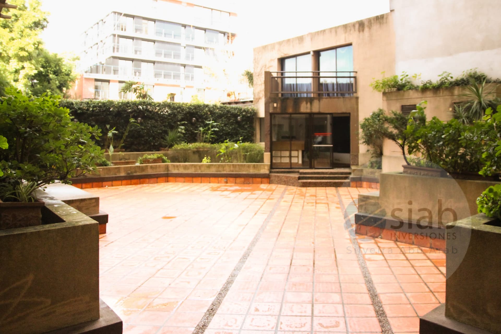 This is an exterior courtyard or terrace area paved with terracotta tiles and a central gravel inlay strip, bordered by raised concrete planters filled with mature shrubs, palms, and ornamental greenery. A modern two-story building with large glass windows, a small balcony with a metal railing, and a glass entry door with stone steps anchors the space, with a multi-story residential building visible beyond the hedge line.
WATERMARK: siab Inversiones