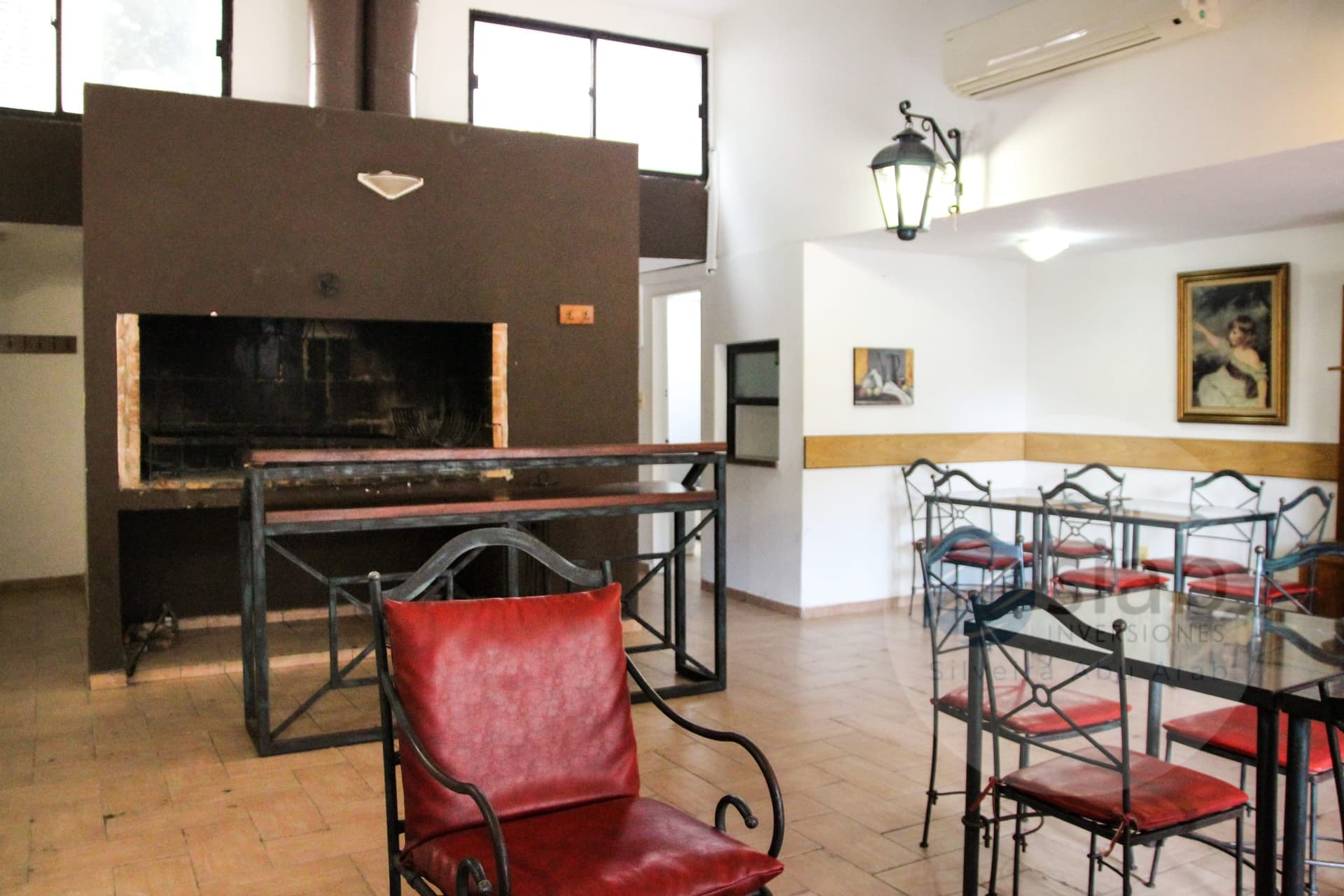 This interior space is a communal dining and gathering area featuring a large dark-brown masonry barbecue (parrilla) with an open firebox and chimney flue, plus a long wrought-iron table with wooden top. Furnishings include red leather-upholstered wrought-iron armchairs, glass-top dining tables with matching red-cushioned chairs, and a wall-mounted air conditioning unit, decorative wall lantern, and framed artwork. The tiled terracotta-style floor and wood wainscoting give it a rustic, well-maintained feel with good natural light from the large windows.
WATERMARK: