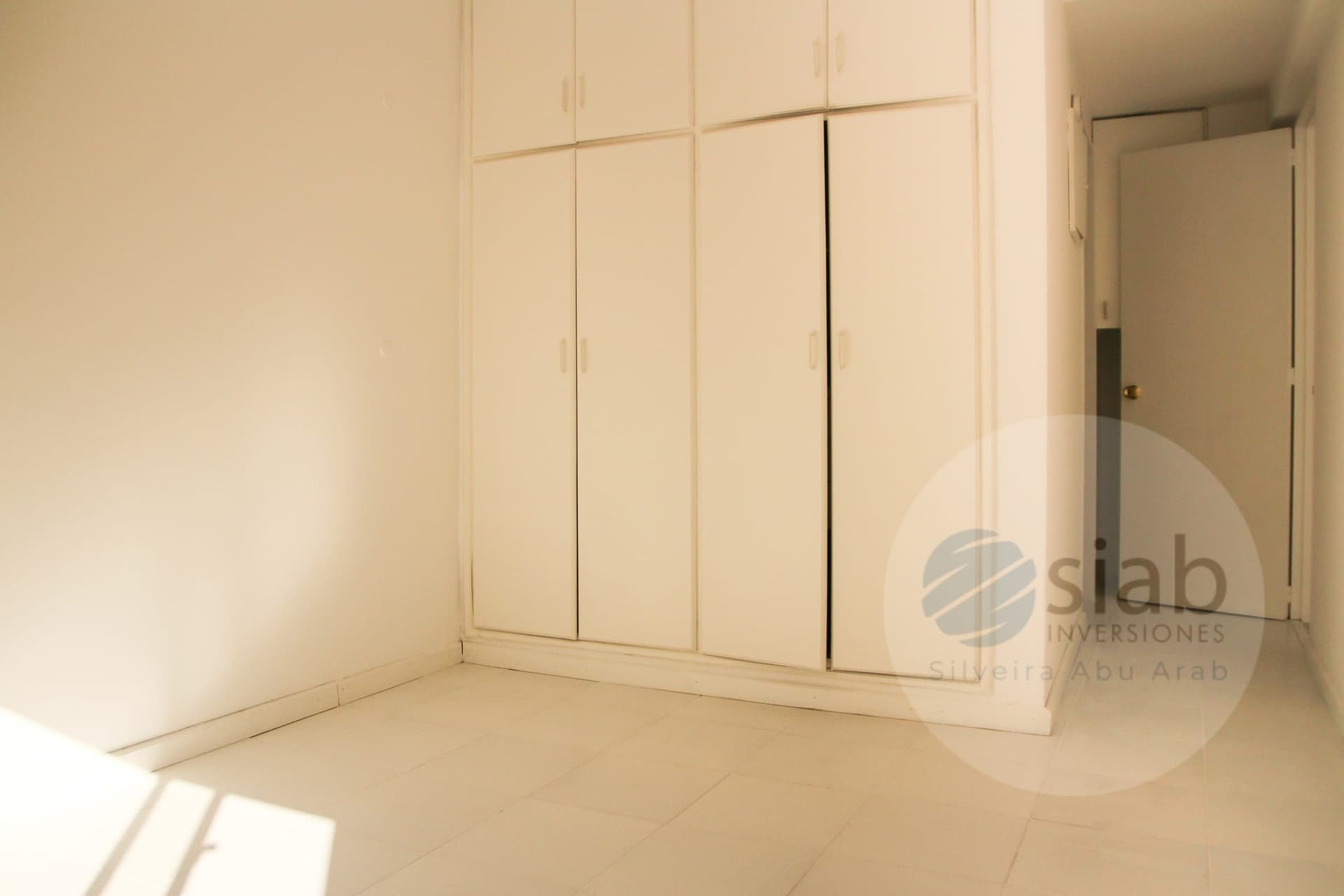 This is an empty bedroom with white-painted walls and a tiled floor in a light cream tone. A full-height built-in wardrobe with eight white panel doors and recessed handles runs along the back wall, and sunlight streams in from the left, casting bright patches across the floor. The room appears clean and unfurnished, with an open doorway on the right leading to a hallway.
WATERMARK: siab Inversiones Silveira Abu Arab