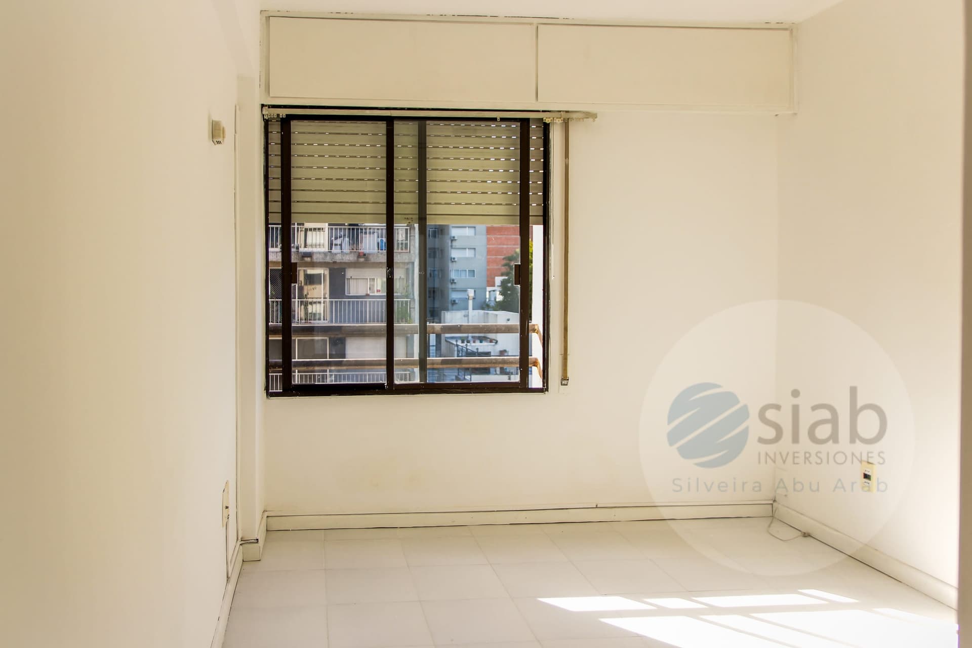 This is an empty bedroom (or similar interior room) with off-white walls and a white tiled floor. A large dark-framed sliding window with a roll-down shutter looks out onto neighboring apartment buildings with balconies, and sunlight casts bright patterns across the floor. The space appears clean and unfurnished, with a wall-mounted fixture and electrical outlets visible.
WATERMARK: Siab Inversiones (Silveira Abu Arab)