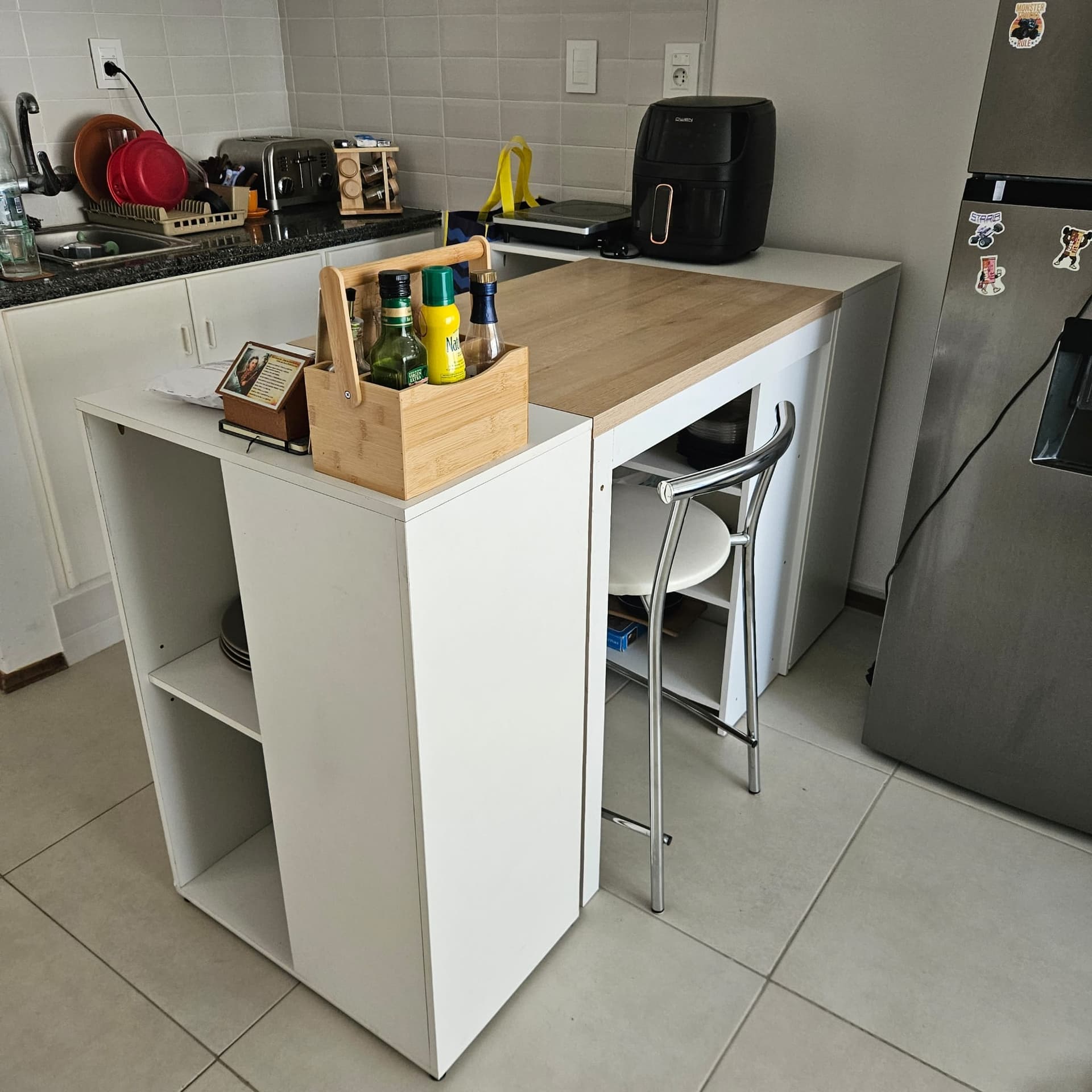 This image shows a compact kitchen with white tiled walls, a granite countertop with an undermount sink, and white base cabinets. In the foreground is a white freestanding bar/island unit with an oak-finished top, open shelving, and a chrome-legged stool tucked underneath, alongside a stainless steel refrigerator and a black air fryer on the counter. The space appears lived-in and in good condition, with pale tiled floors and no visible watermark or agency logo.