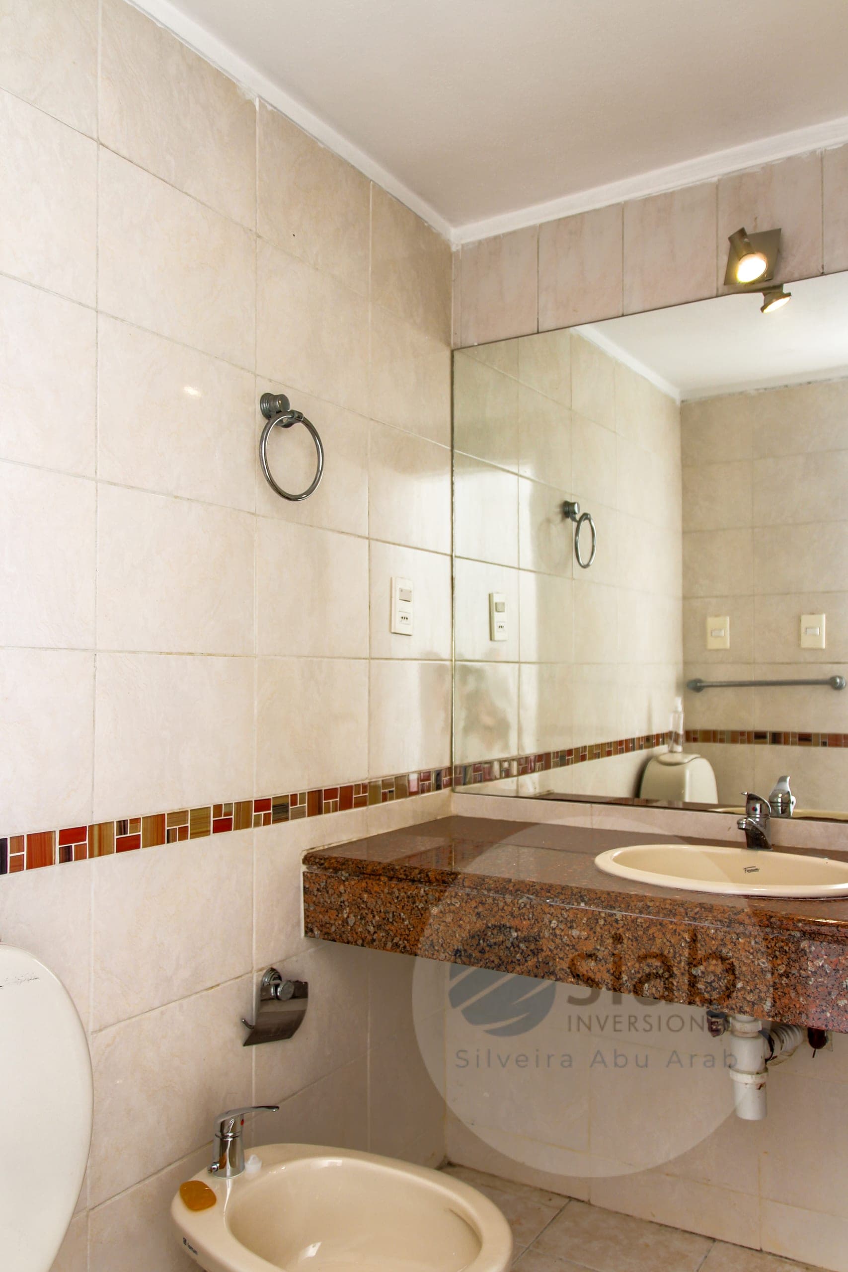 This is a bathroom featuring cream-toned wall tiles, a decorative mosaic accent strip in red and brown tones, and a granite vanity with an inset cream oval sink and chrome faucet. A large wall mirror, chrome towel ring, toilet paper holder, and spotlights complete the space, with a bidet visible in the foreground. The fixtures appear functional and the room looks somewhat dated but generally well maintained.
WATERMARK: Stiabo Inversiones (Silveira Abu Arab)