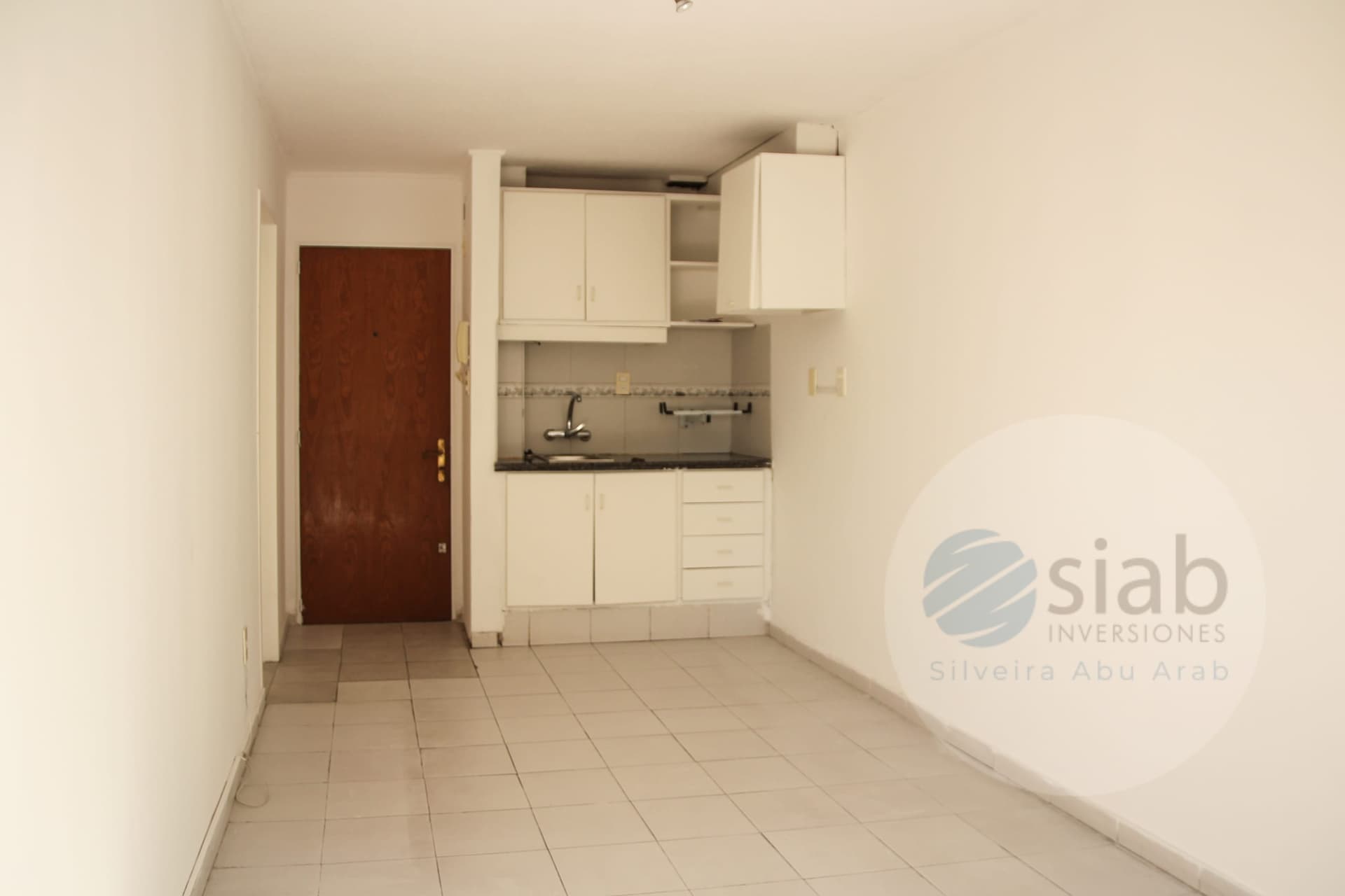 This image shows a small, empty studio-style living area or compact kitchen space with light beige walls and large beige ceramic floor tiles. The compact kitchenette has white upper and lower cabinets, a four-drawer unit, a dark granite-look countertop, and a single chrome faucet over a stainless sink, with a tiled backsplash. The room is unfurnished and appears clean but dated, with a wooden entry door featuring a gold-toned lock and an intercom panel beside it.
WATERMARK: SIAB Inversiones (Silveira Abu Arab)